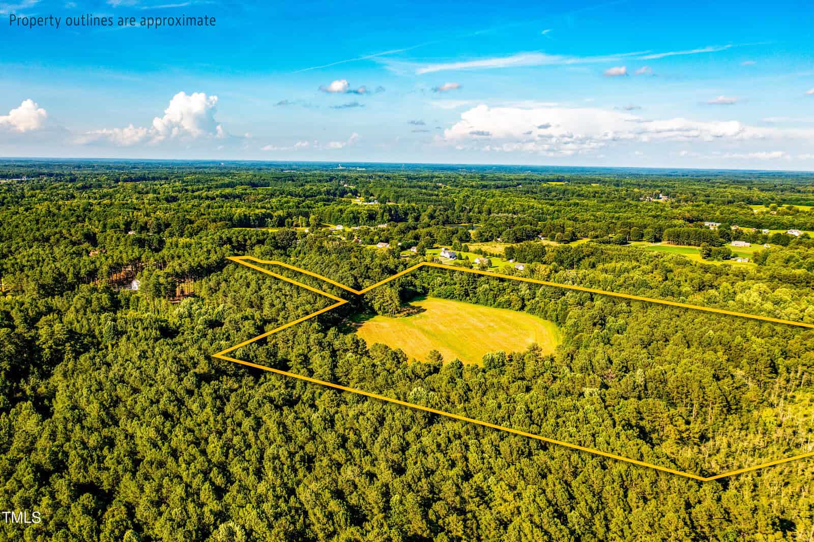 Aerial view of a forested landscape with a large open grassy field in the center, outlined with yellow lines to indicate approximate property boundaries. Blue sky with clouds is visible above the green scenery.