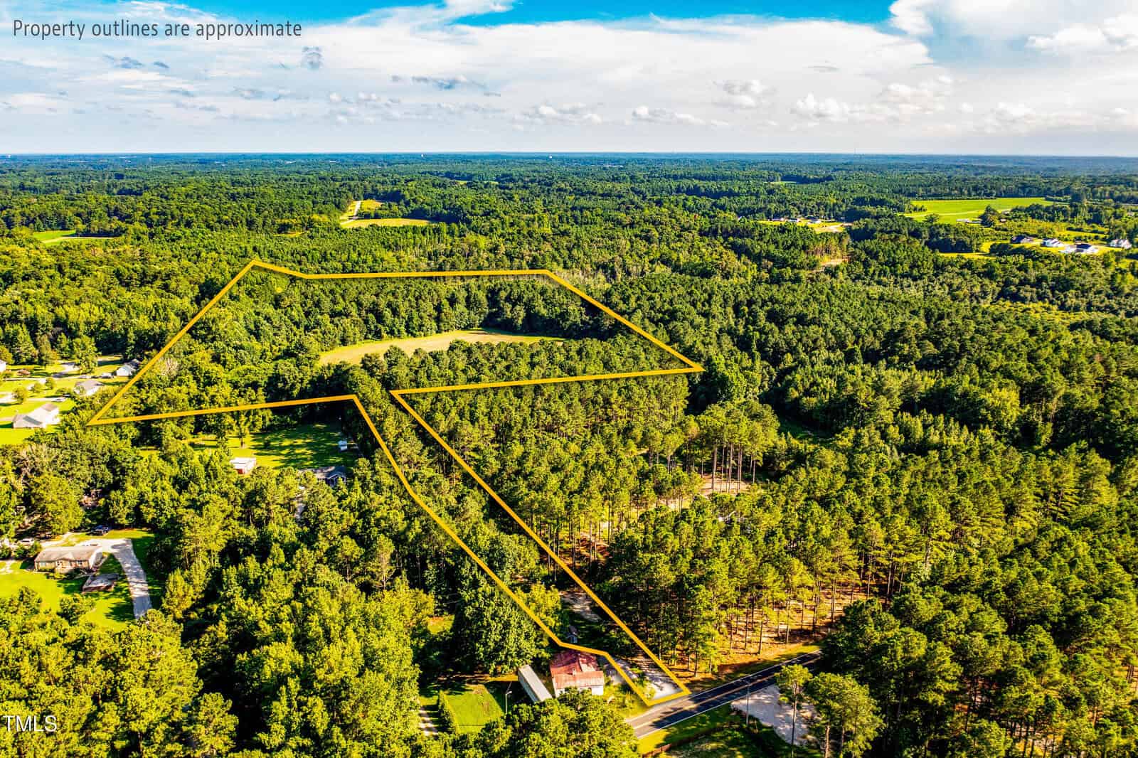 Aerial view of a wooded landscape with the approximate property boundary outlined in yellow, encompassing a large area of forest, some open space, and a few nearby houses; blue sky with scattered clouds above.
