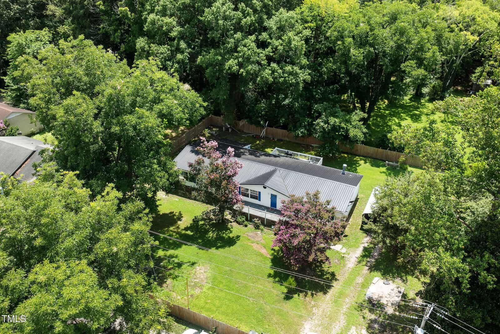 Aerial view of a single-story house with a dark roof, surrounded by trees and a fenced grassy yard. A gravel driveway leads up to the home, and flowering trees are in the front yard. Dense woods border the backyard.