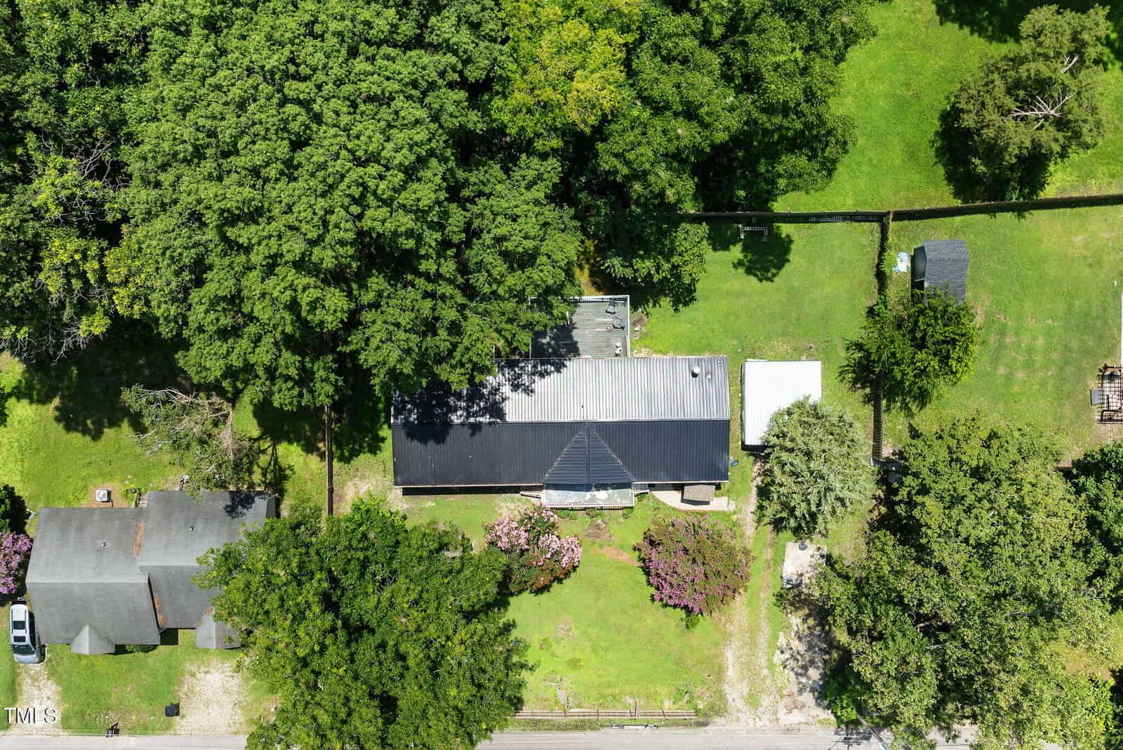 Aerial view of a house with a dark roof surrounded by trees and a grassy yard; a driveway and neighboring buildings are visible, with a road at the bottom of the image.