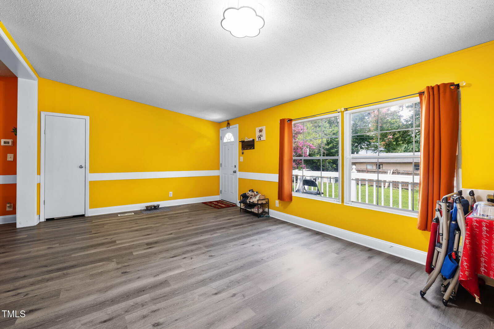 Bright living room with yellow walls, white trim, large front window, wood-look flooring, and a small pet crate by the door. Curtains frame the window, and various household items are visible along the walls.
