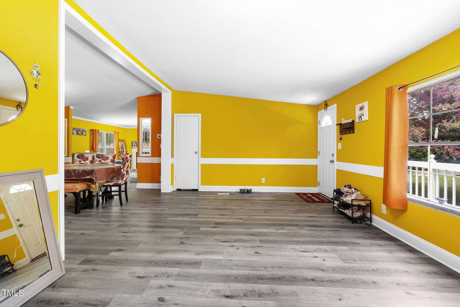 Bright living room with bold yellow walls, white trim, and gray wood flooring. Large window with orange curtain lets in natural light. Dining area with table is visible through a wide doorway.