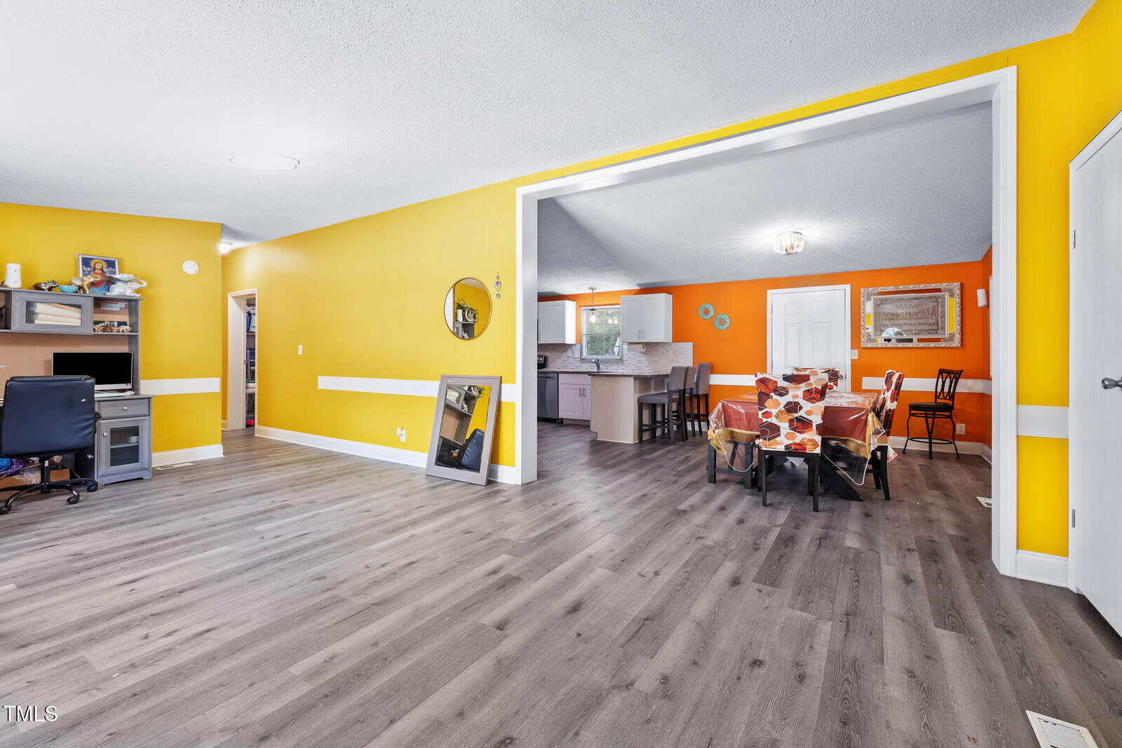 Bright open-plan room with yellow and orange walls, wood flooring, a dining area with a table and chairs, a small kitchen, desk with office chair, artwork, and white doors. Natural light brightens the space.