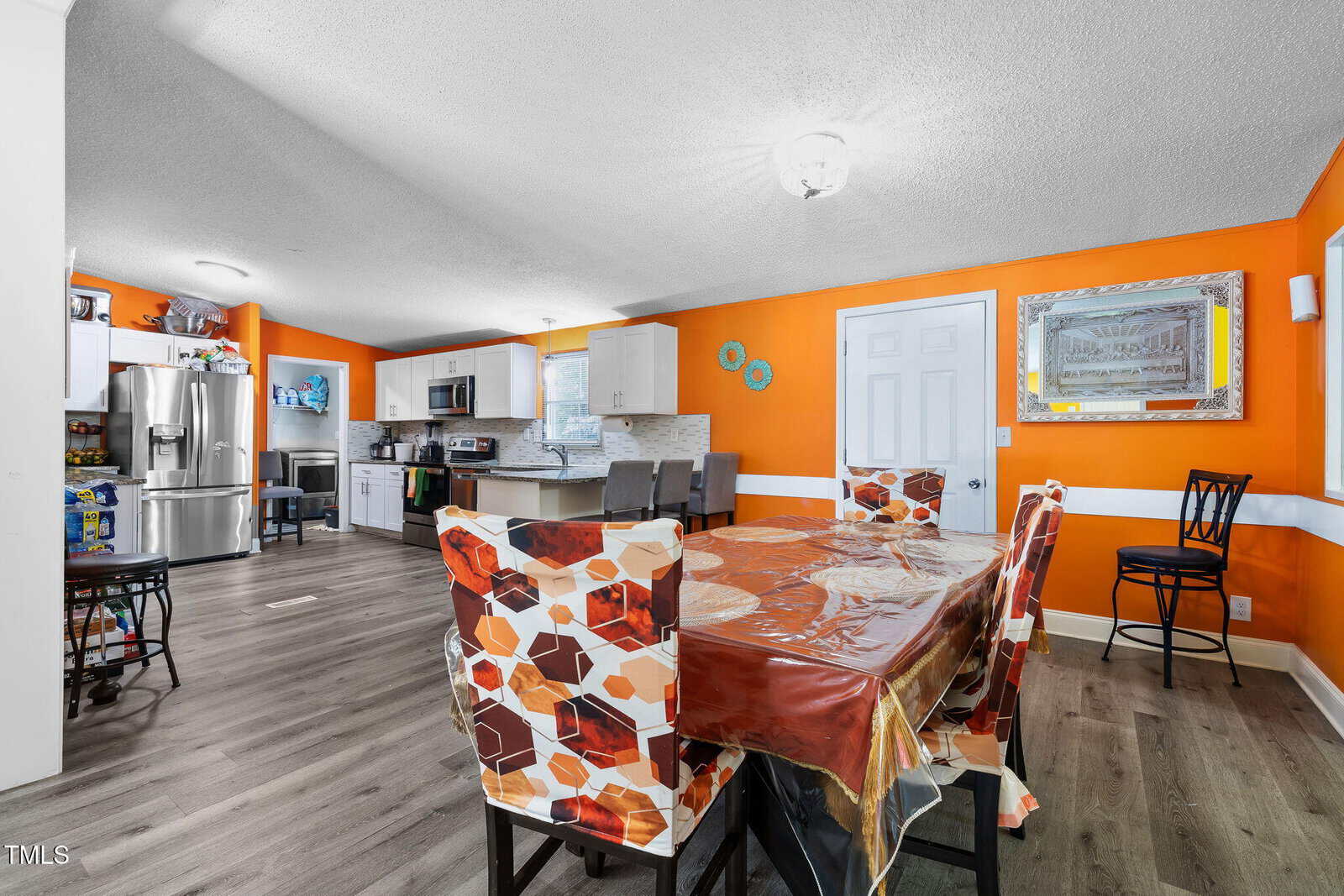 Bright kitchen and dining area with orange walls, patterned chairs, and a table covered with a plastic tablecloth. Kitchen features stainless steel appliances, white cabinets, and a breakfast bar with stools.