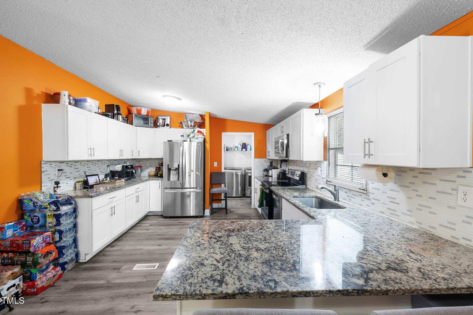 Modern kitchen with bright orange walls, white cabinets, stainless steel appliances, granite countertops, and stacked pantry items. Sunlight streams through a window above the sink, illuminating the clean, organized space.