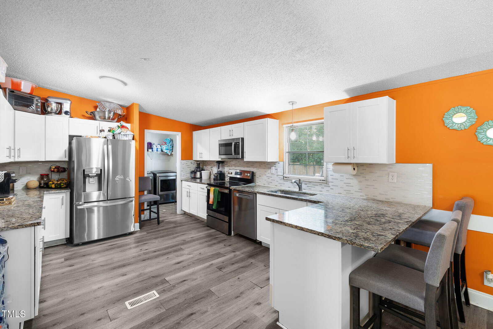Modern kitchen with bright orange walls, white cabinets, granite countertops, stainless steel appliances, and light wood flooring. There is a breakfast bar with gray stools and decorative wall accents. Sunlight streams in through a window.