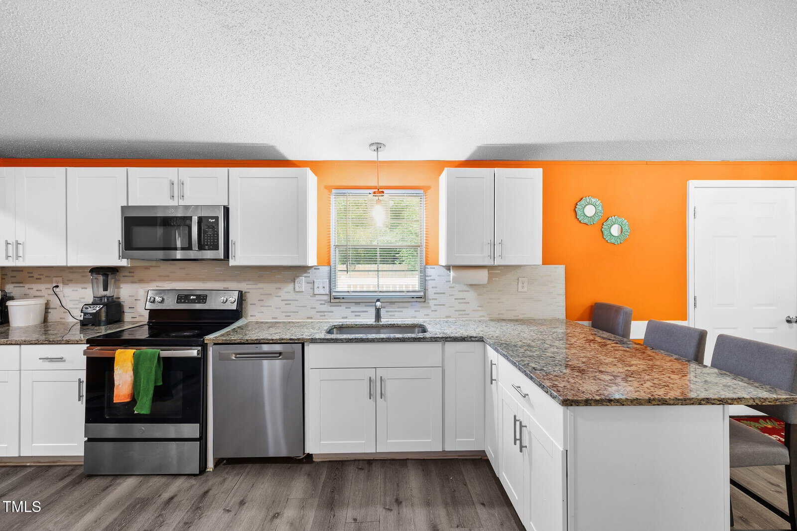 Bright kitchen with white cabinets, stainless steel appliances, granite countertops, orange accent wall, and a window above the sink. Four gray chairs line the breakfast bar. Two green decorative plates hang on the wall.