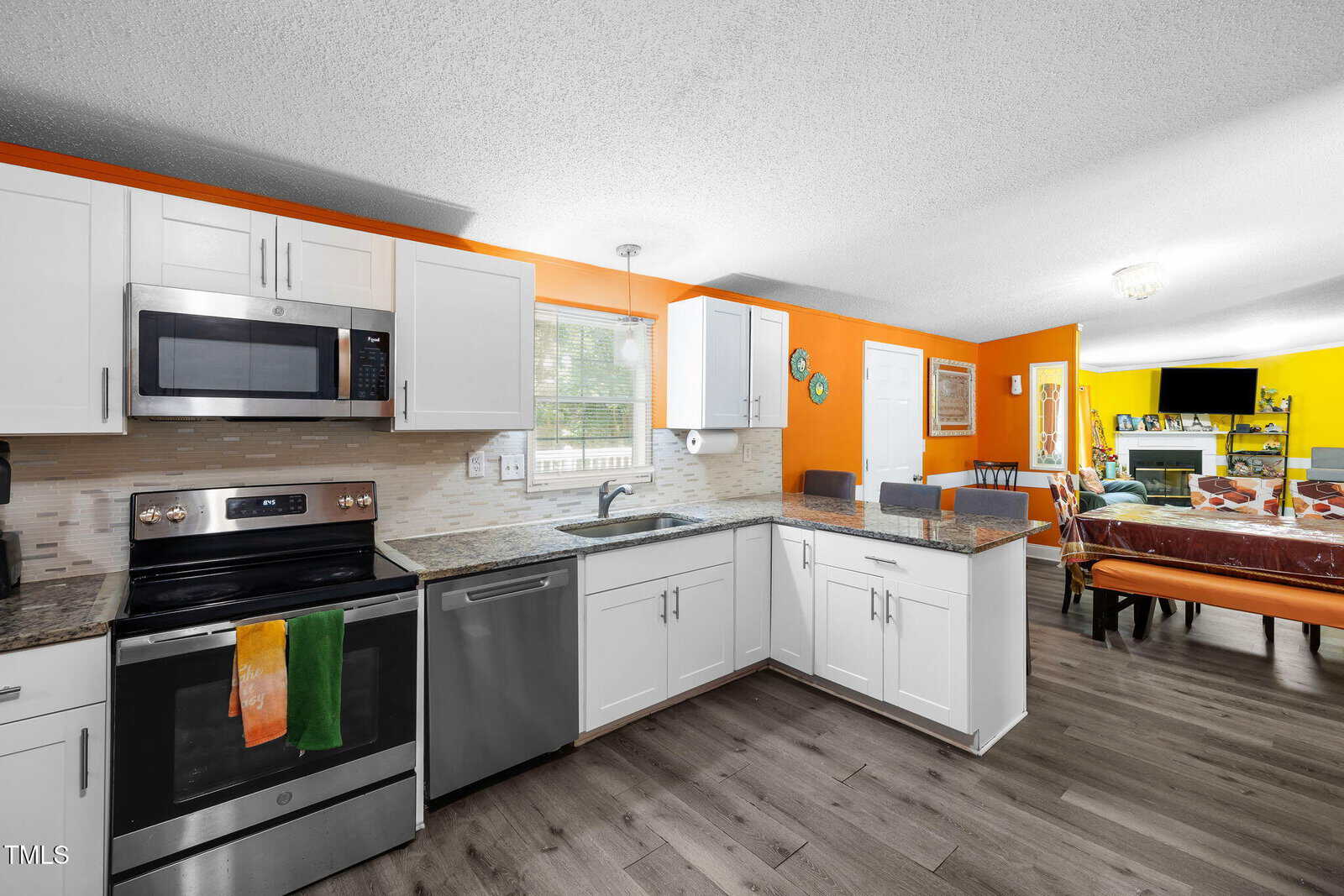 Modern kitchen with white cabinets, stainless steel appliances, and a bright orange accent wall. The space opens to a dining area with a table, chairs, and a living room with a TV and yellow accent wall.
