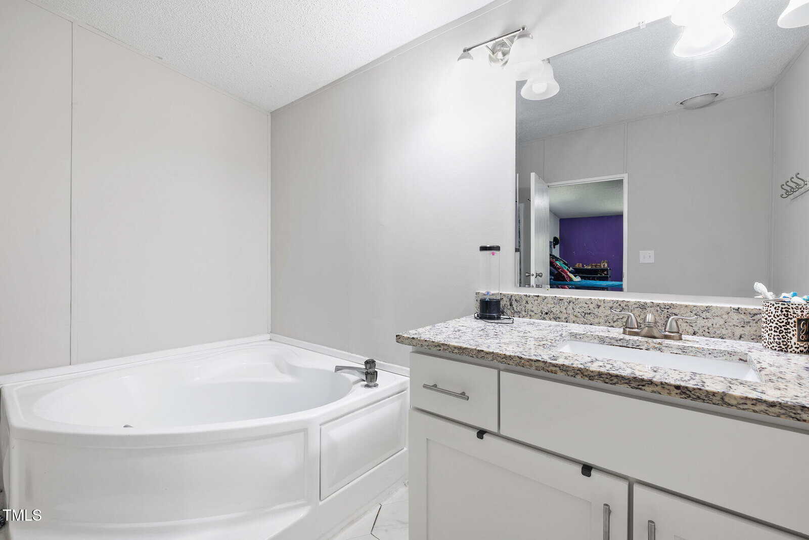 Modern bathroom with a white corner bathtub, granite countertop vanity with dual sinks, and a large mirror. The room is well-lit and has white cabinets and light-colored walls.