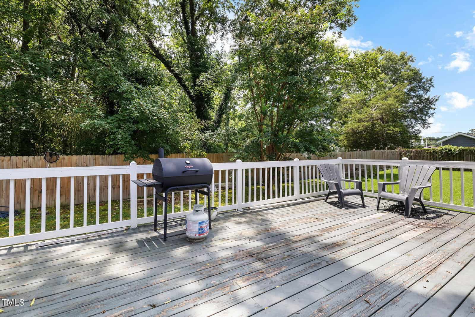 A backyard wooden deck with a white railing features a black grill and two gray Adirondack chairs. Trees and a wooden fence surround the green lawn in the background under a partly cloudy sky.