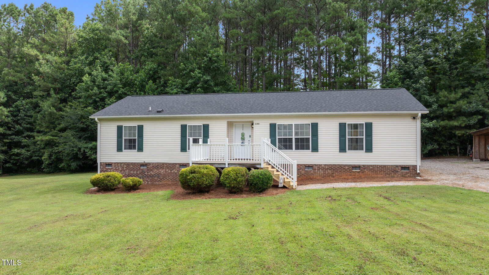 A single-story house with white siding, dark shutters, and a brick foundation. A small front porch with stairs faces a well-kept lawn, surrounded by shrubs and bordered by dense trees.