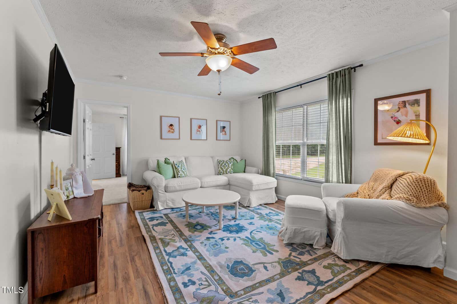 Bright living room with a ceiling fan, white armchairs and sofa, blue and green patterned rug, round white coffee table, wall art, floor lamp, and large window with green curtains letting in natural light.
