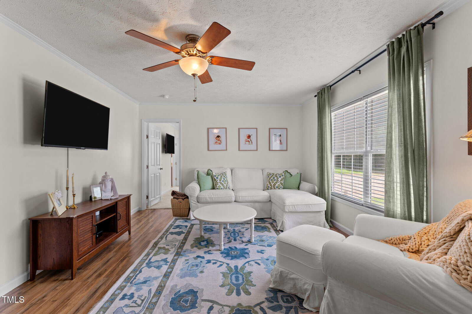 Bright living room with white walls, a ceiling fan, large window with green curtains, white sectional sofa, ottoman, patterned rug, wooden TV stand, and framed art on the wall.