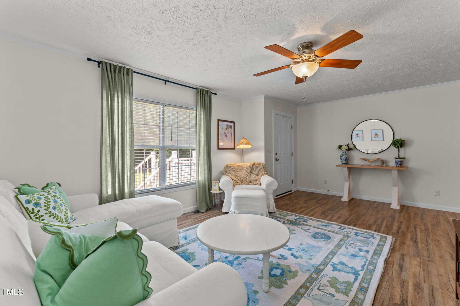 A bright living room with a white sofa, green and blue pillows, and a matching armchair. A round coffee table sits on a floral rug, and a console table with a round mirror is against the far wall. Green curtains frame a large window.