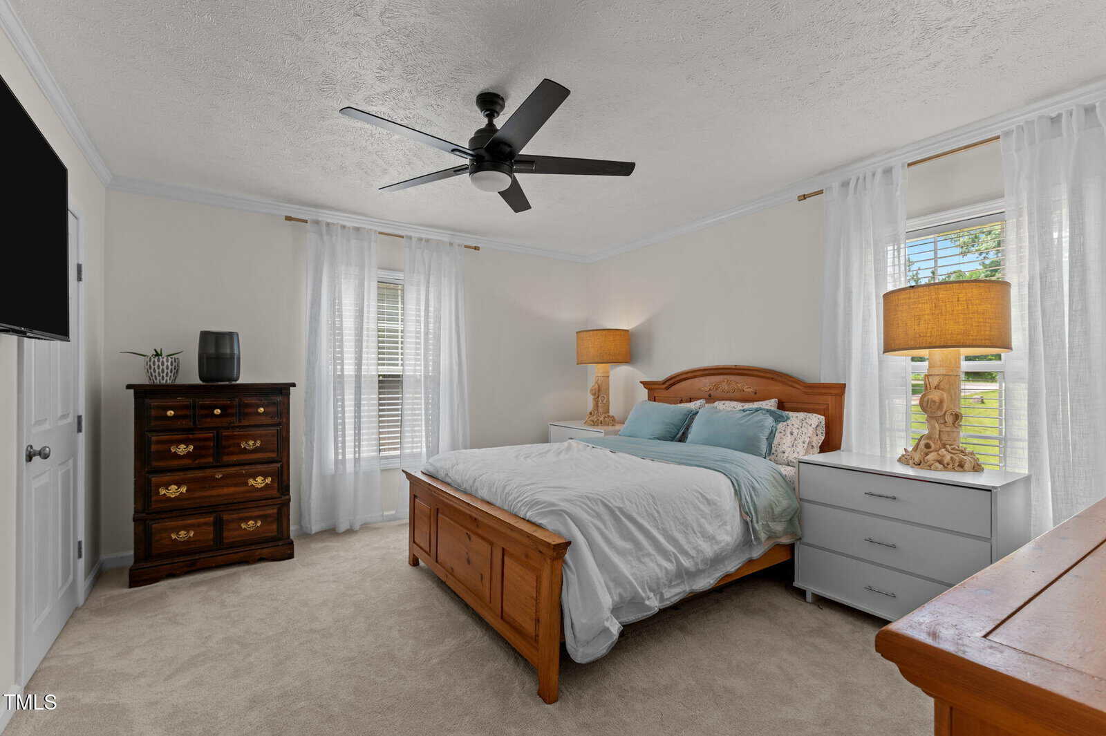 A bedroom with a wooden bed, white bedding, two nightstands with lamps, a dark dresser, a ceiling fan, beige carpet, and white curtains covering windows.