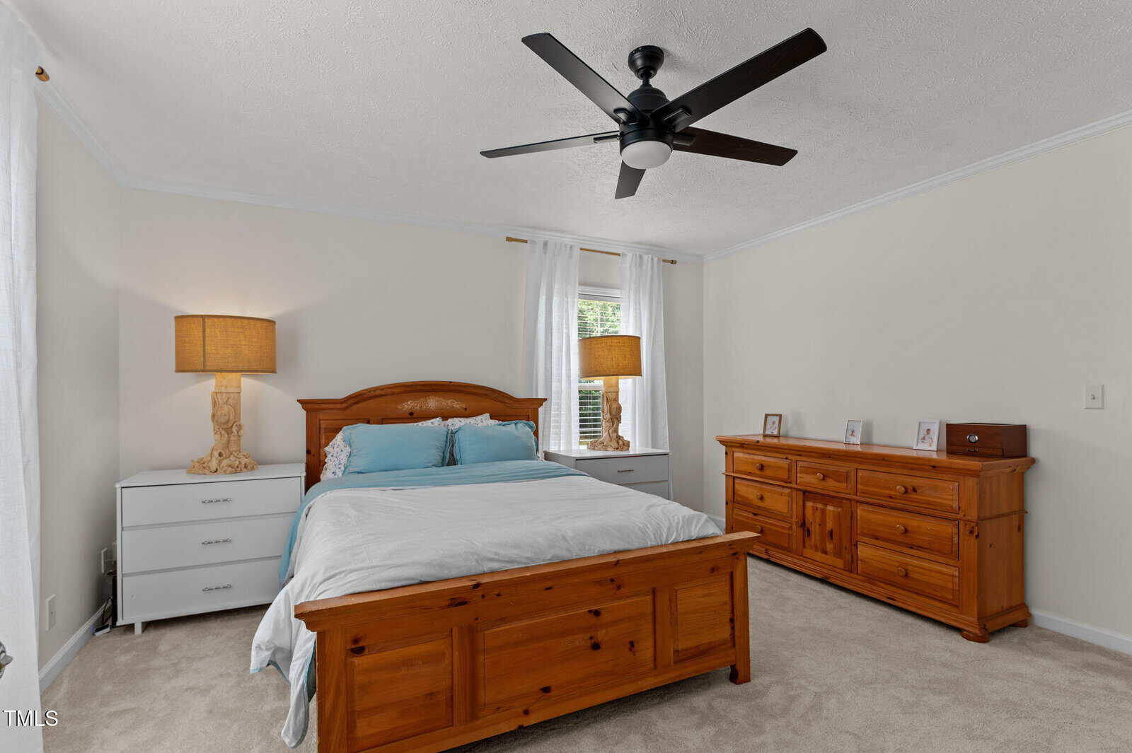 A bedroom with a wooden bed frame, matching dresser and nightstands, two table lamps, white bedding, beige carpet, white walls, a ceiling fan, and a window with sheer white curtains.
