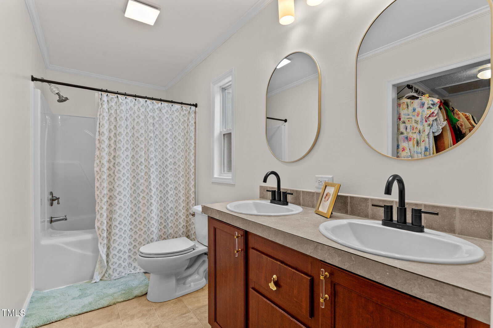 Bathroom with double sinks, dark faucets, a large wooden cabinet, a toilet, and a bathtub with a patterned shower curtain. A window is above the toilet; a closet with clothes is visible in a mirror reflection.