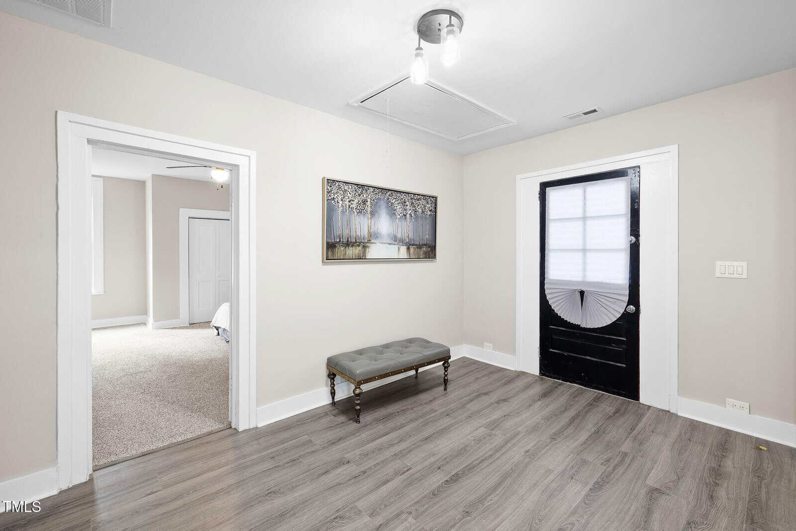 A small, empty room with light gray walls, wood flooring, a black door with a window shade, a wall bench, a framed painting, and an open doorway leading to a carpeted bedroom.