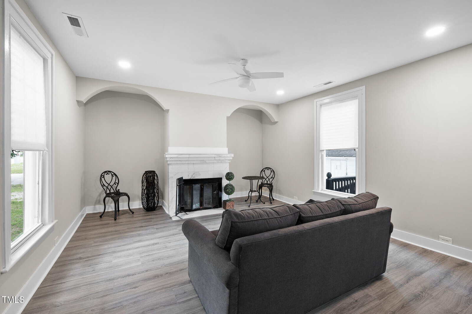 A living room with light gray walls, wood flooring, a dark gray sofa, a white fireplace, and two small round tables with chairs by the walls. Large windows bring in natural light.