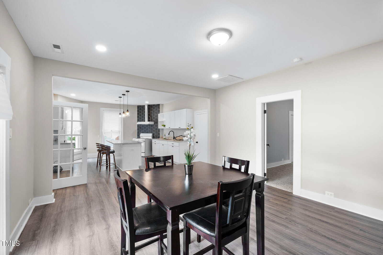 A modern dining area with a dark wood table and six chairs sits next to an open kitchen featuring white cabinets, a center island, and stainless steel appliances. The floor is wood and walls are light-colored.
