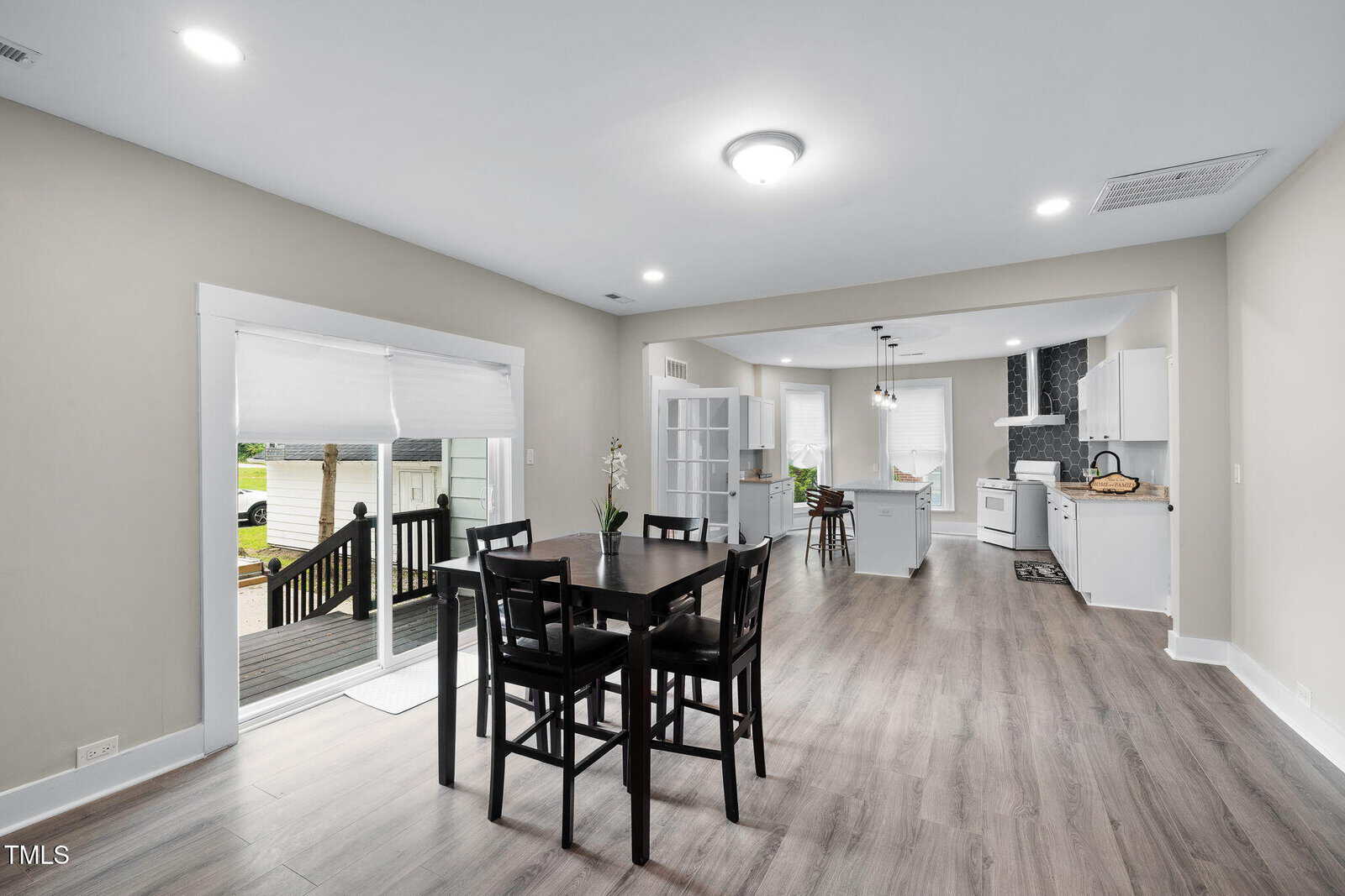 A dining area with a dark wood table and four chairs sits on wood flooring between a sliding glass door and an open kitchen with white cabinets, an island, and stainless steel appliances.