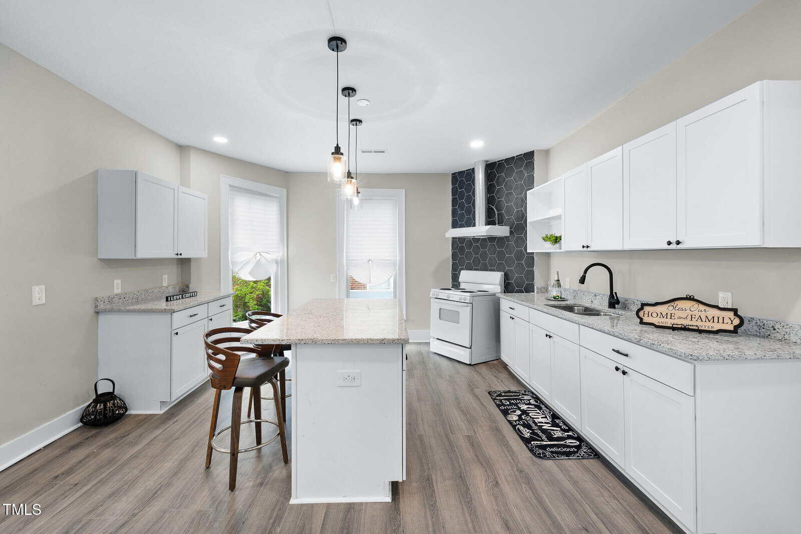Modern kitchen with white cabinets, granite countertops, a central island with barstools, wood flooring, pendant lights, gas stove, hexagon tile backsplash, and large windows with white shades.