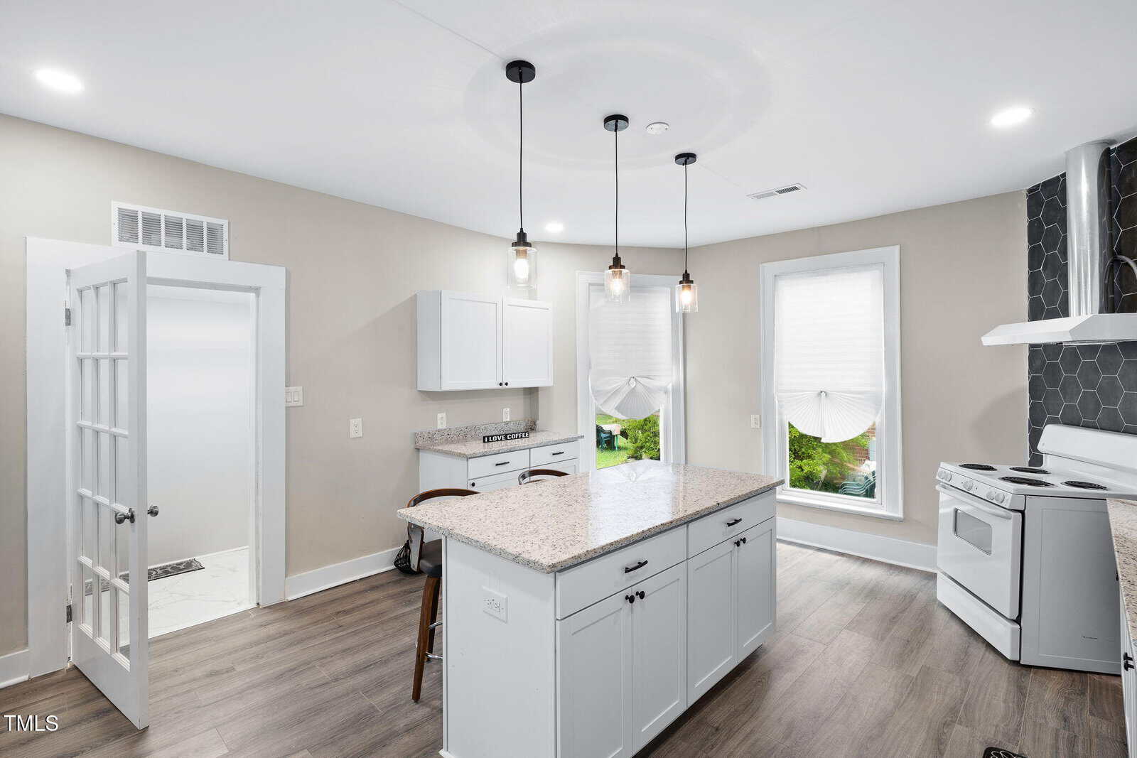 Modern kitchen with white cabinets, granite countertops, pendant lights, and wood flooring. Large windows with shades let in natural light. Stools sit at the island, and a stove is against a hexagon-tiled backsplash.