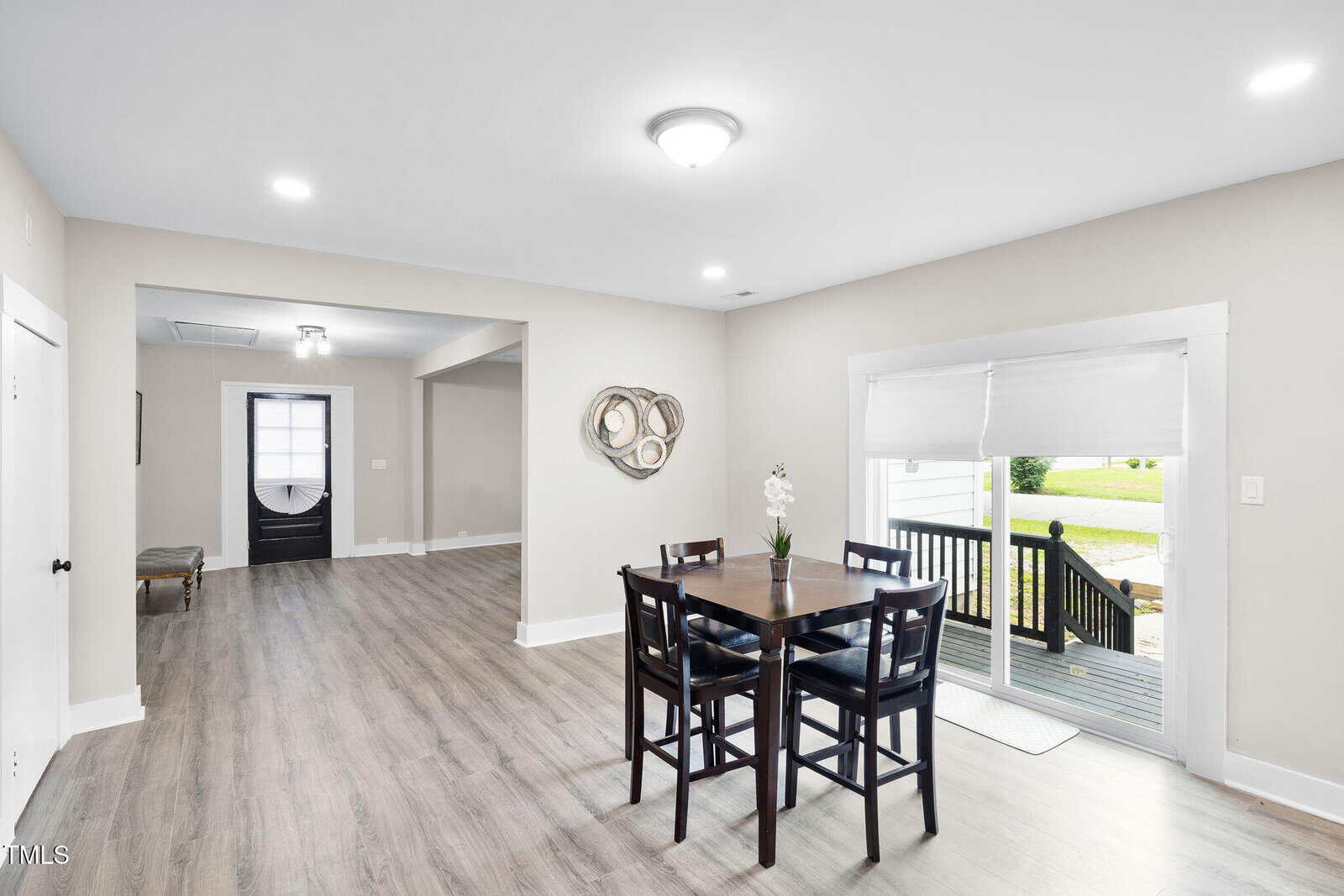 Bright dining area with a dark wood table and four chairs, light wood floors, and adjacent sliding door leading outside. Open layout connects to a hallway with visible front door and decorative wall art.
