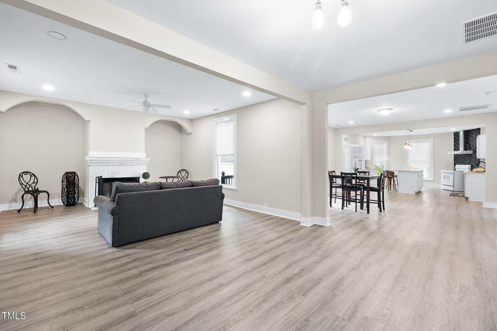Spacious open-plan living area with light wood flooring, gray sofa, and fireplace, adjacent to a dining area and modern white kitchen with stainless steel appliances; neutral walls and bright lighting throughout.