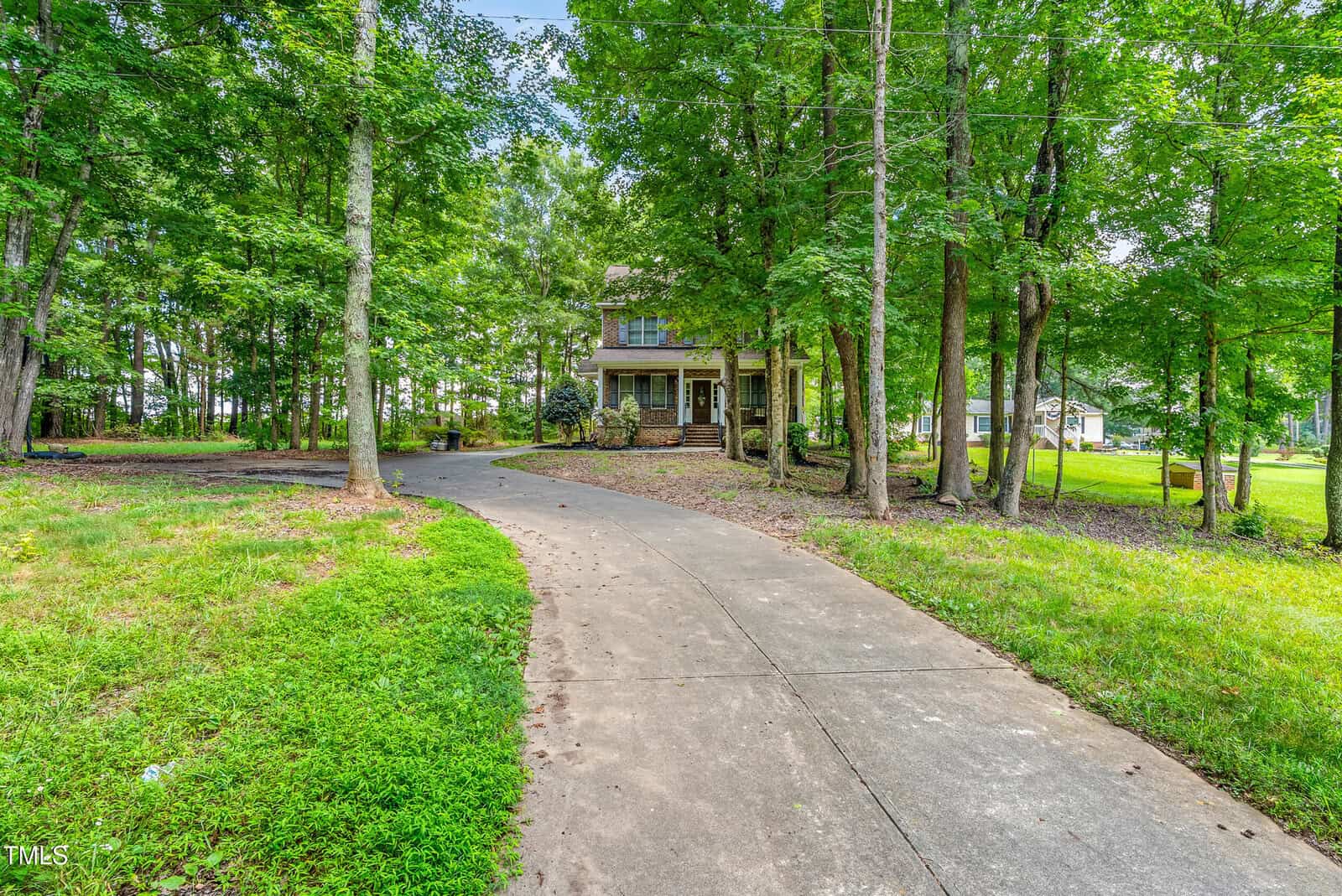 A two-story house with a covered porch is set back from a curved concrete driveway, surrounded by tall trees and green grass. The yard is spacious with patches of mulch and landscaped shrubs near the house.