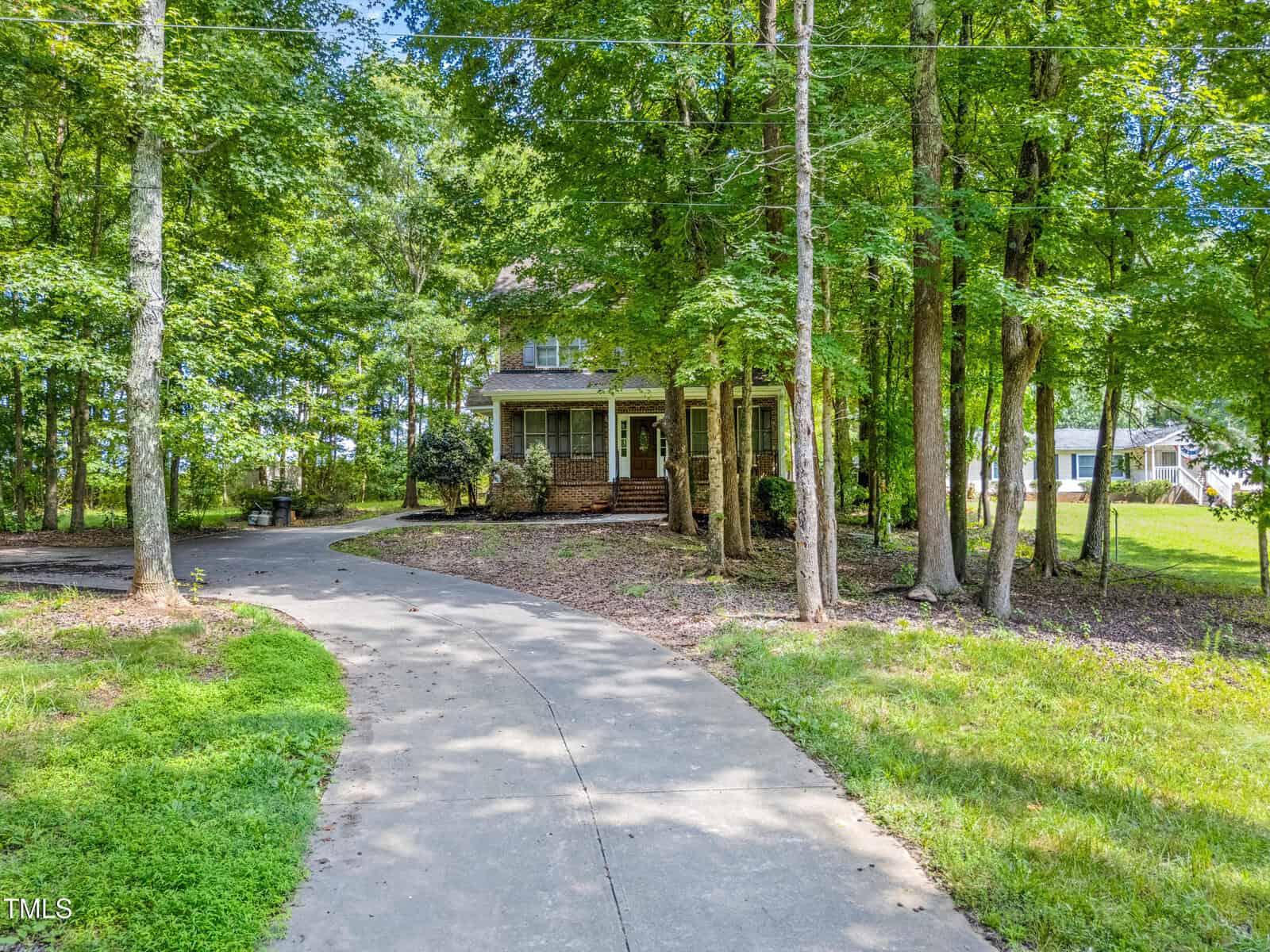 A two-story house with a covered front porch is partially visible behind trees. A curved concrete driveway leads up to the house, surrounded by a grassy yard and shaded by mature trees.