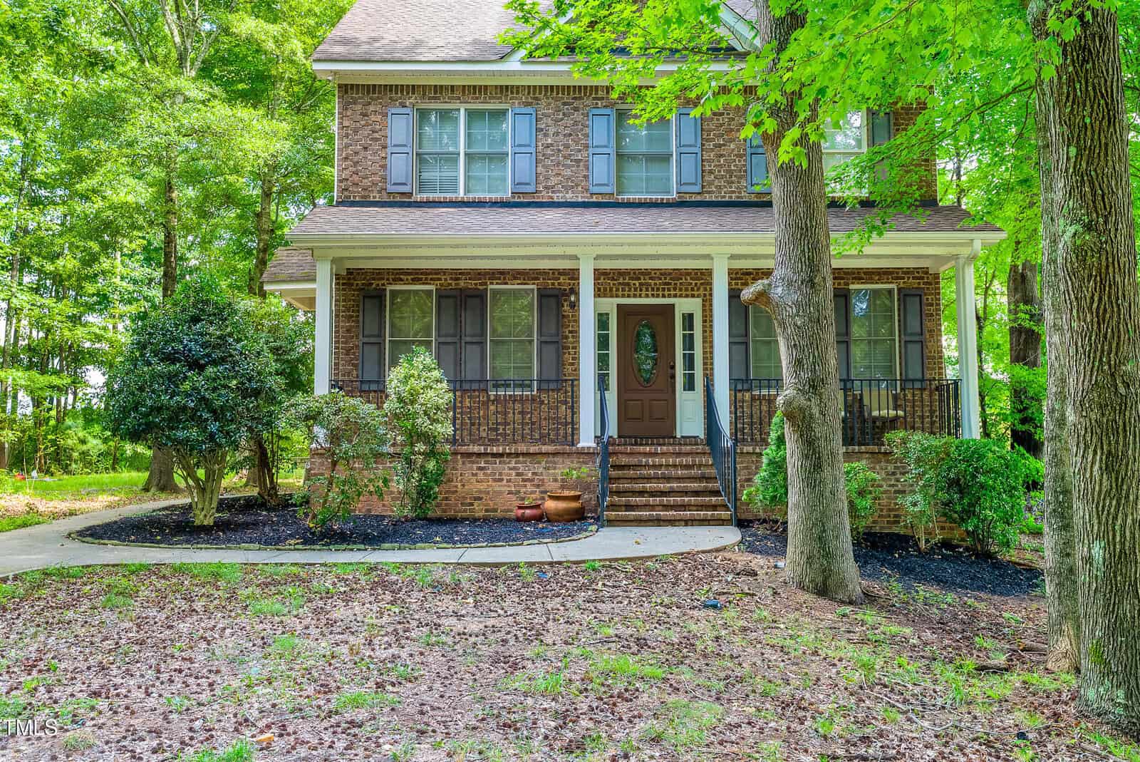 Charming two-story brick house in Franklinton, featuring a covered front porch with black railings and shuttered windows. The inviting brown front door at 4445 W River Road welcomes you home, while the yard boasts mature trees, shrubs, and a curved sidewalk leading to the steps. Leaf litter gently covers parts of the lawn, adding to the property’s natural appeal.