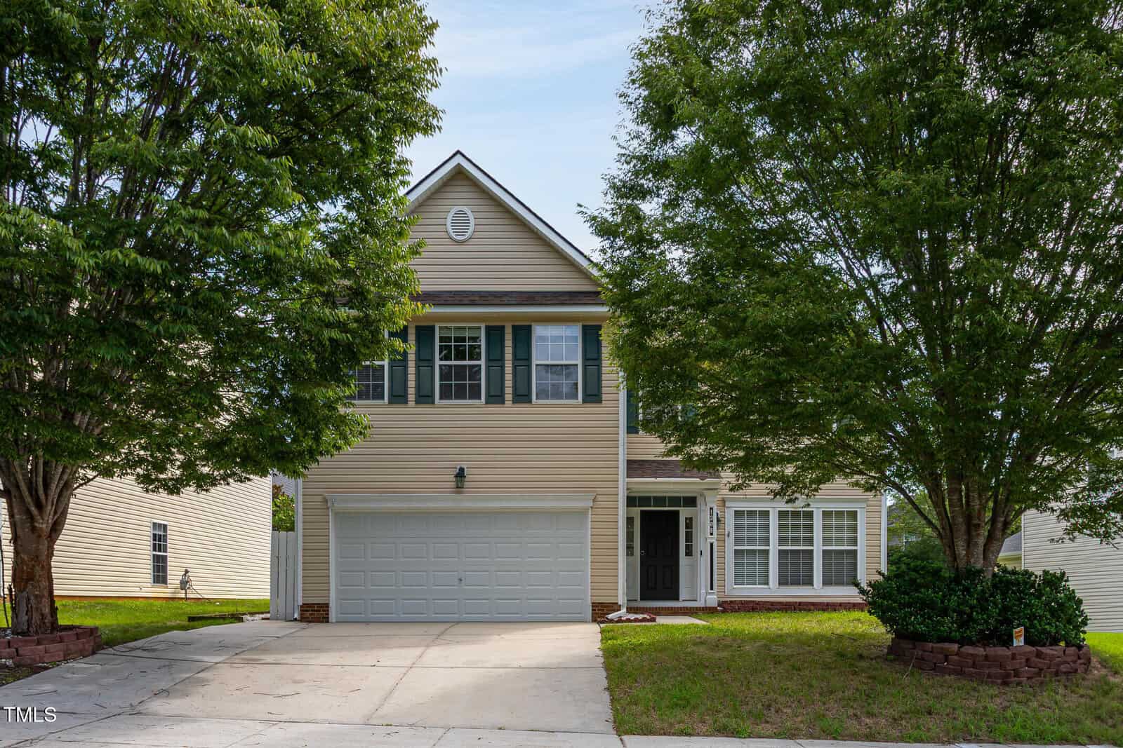 Two-story beige house with white trim and a double garage, partially shaded by large green trees. The front yard has a paved driveway, small shrubs, and a well-kept lawn.