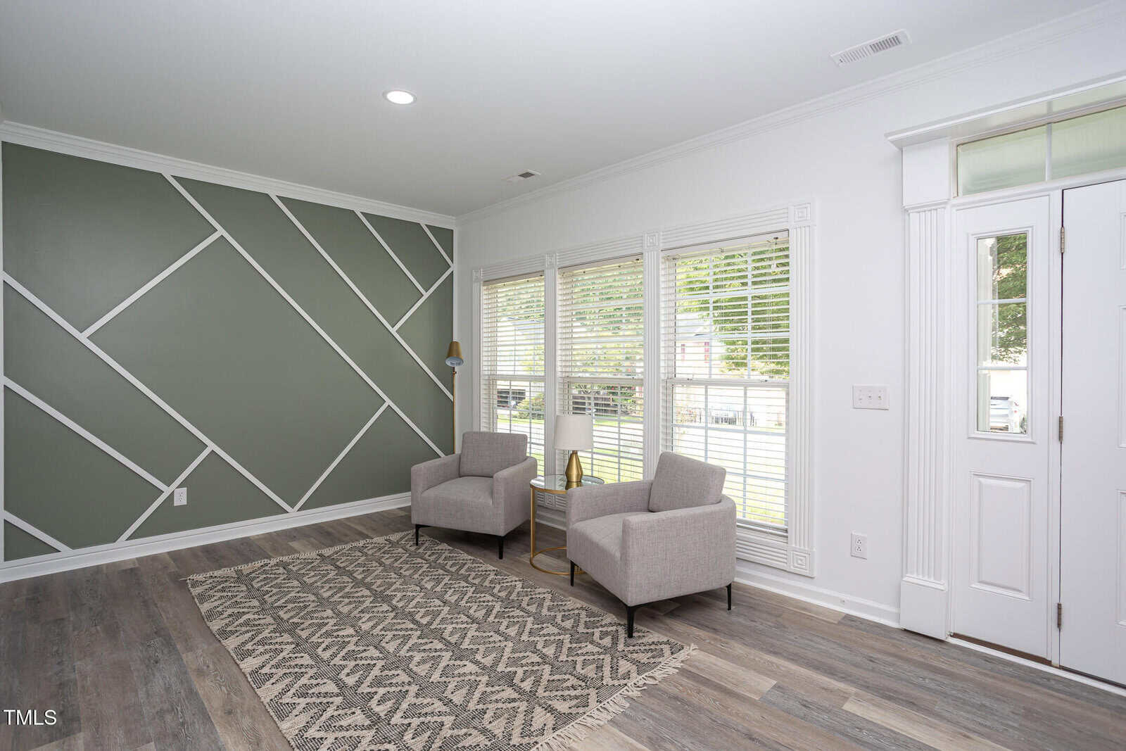 A modern living room with two gray armchairs, a small table, and a patterned rug. One wall features geometric designs in green and white; large windows with blinds let in natural light.