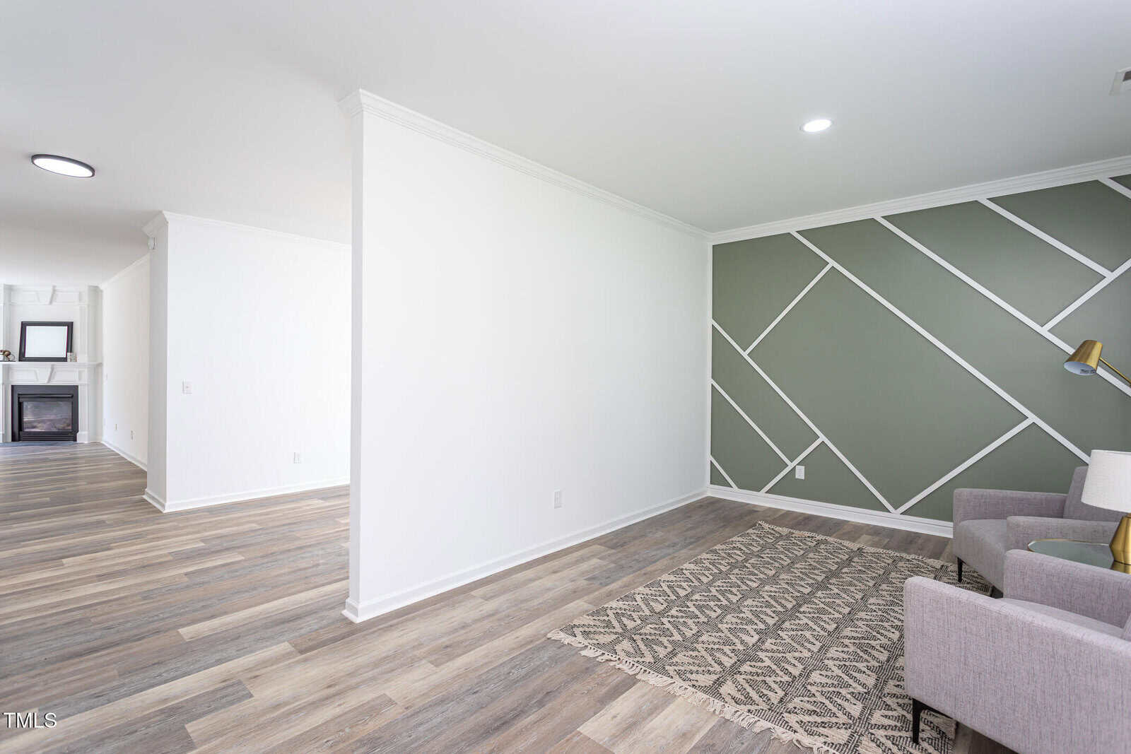 Modern living space with wood-patterned flooring, white walls, and a green accent wall with white geometric lines. Contains a gray armchair, floor lamp, and patterned rug. Fireplace visible in the background.