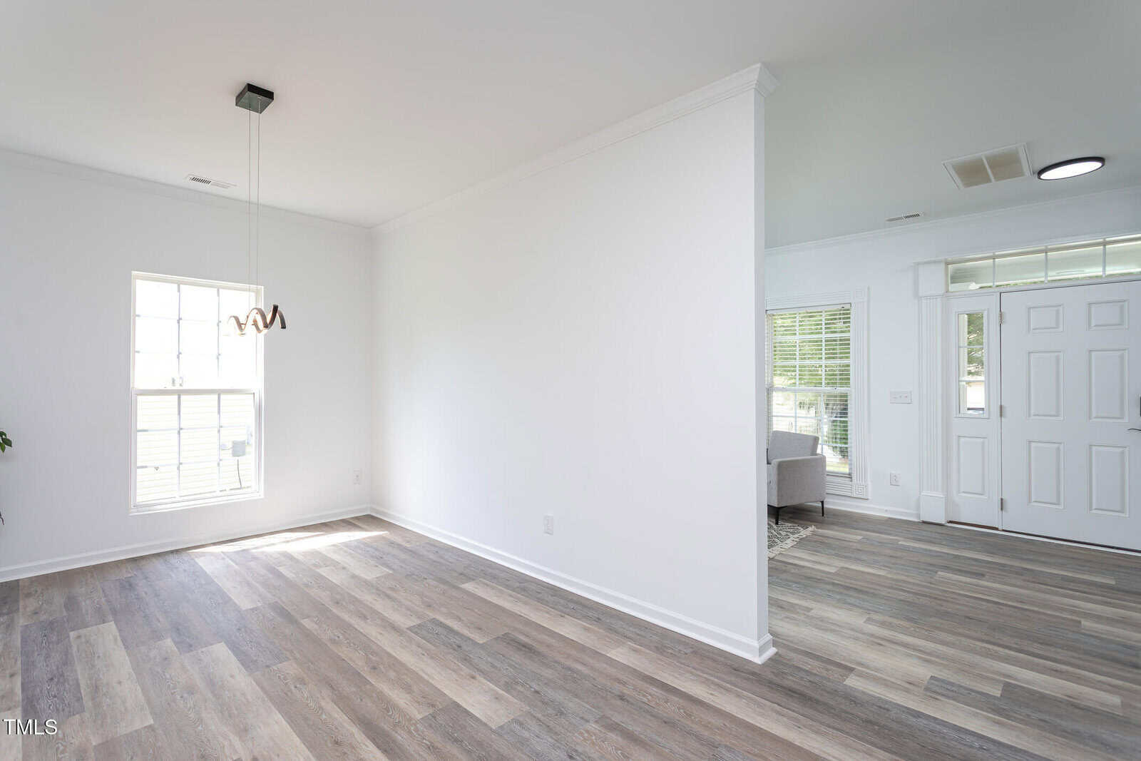 A bright room with light wood flooring, white walls, a modern pendant light, and large windows. A partial wall separates the entryway, which has a white door and more windows, from the rest of the space.