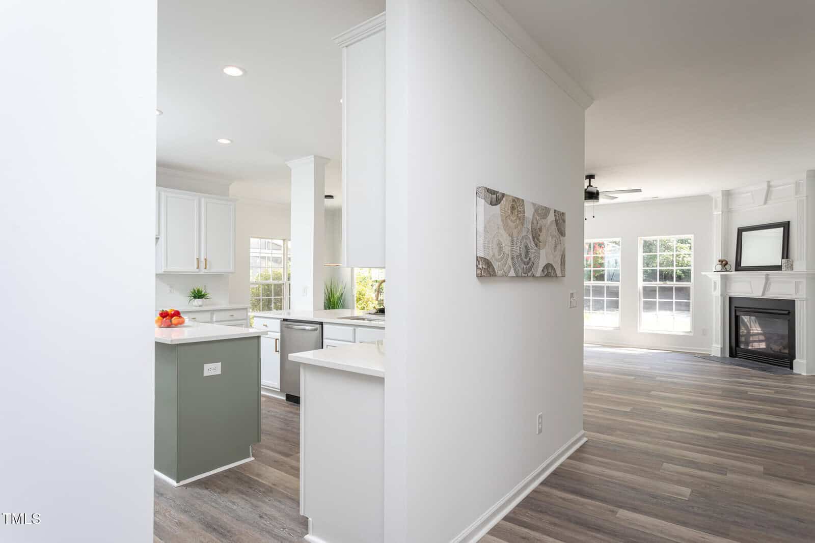 A modern, open-concept kitchen and living area with white walls, light wood floors, white cabinets, and large windows letting in natural light. A decorative painting hangs on the wall, and a fireplace is visible in the living room.