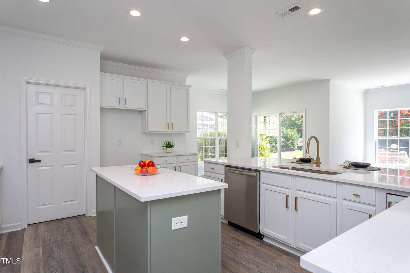 Modern kitchen with white cabinets, light gray island, stainless steel appliances, and a bowl of apples on the island. Large windows let in natural light, and wood flooring runs throughout the space.