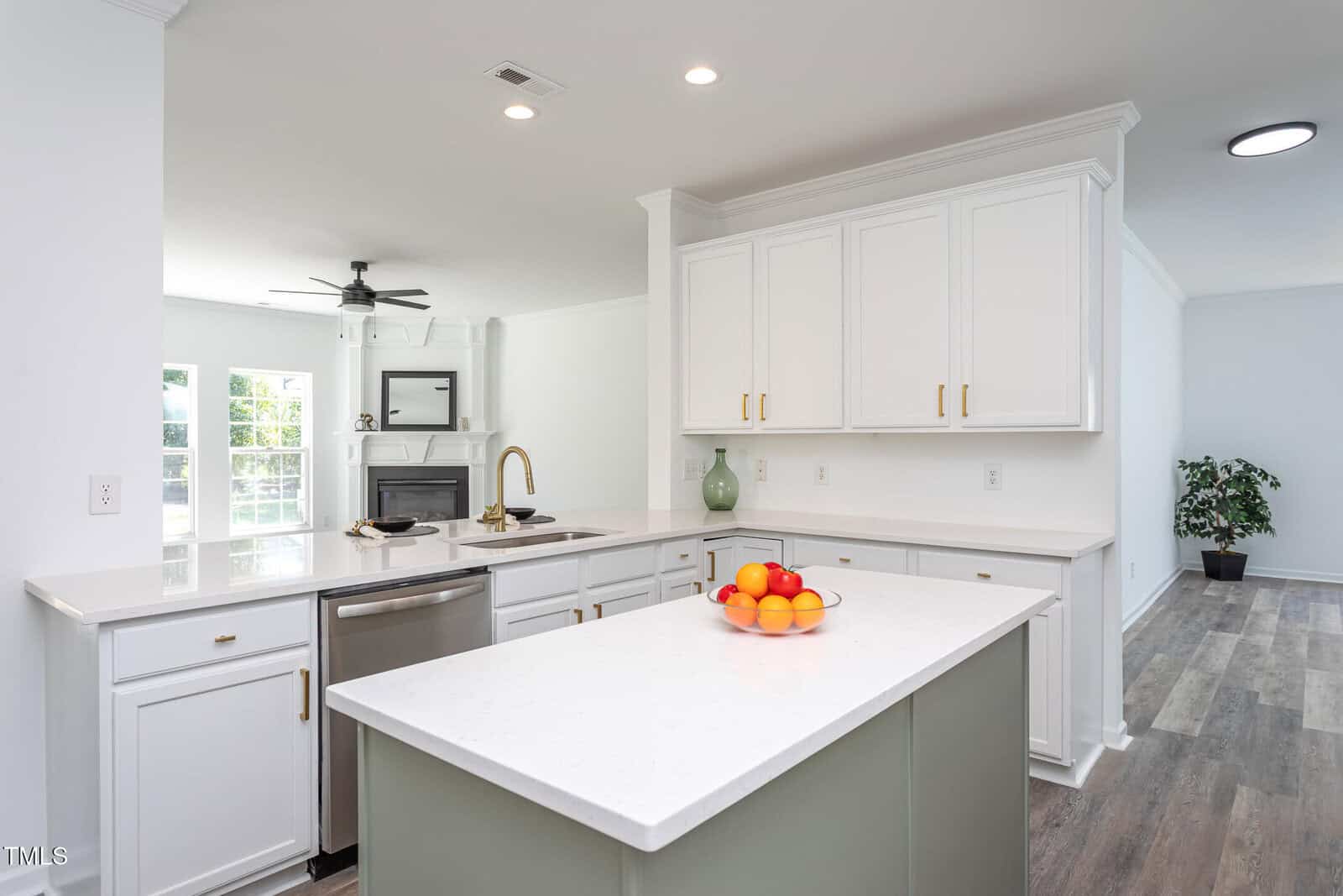 Modern kitchen with white cabinets, gold handles, a stainless steel dishwasher, and a white island featuring a bowl of assorted fruit. The space is well-lit with natural light and has wood flooring. Living room visible in the background.