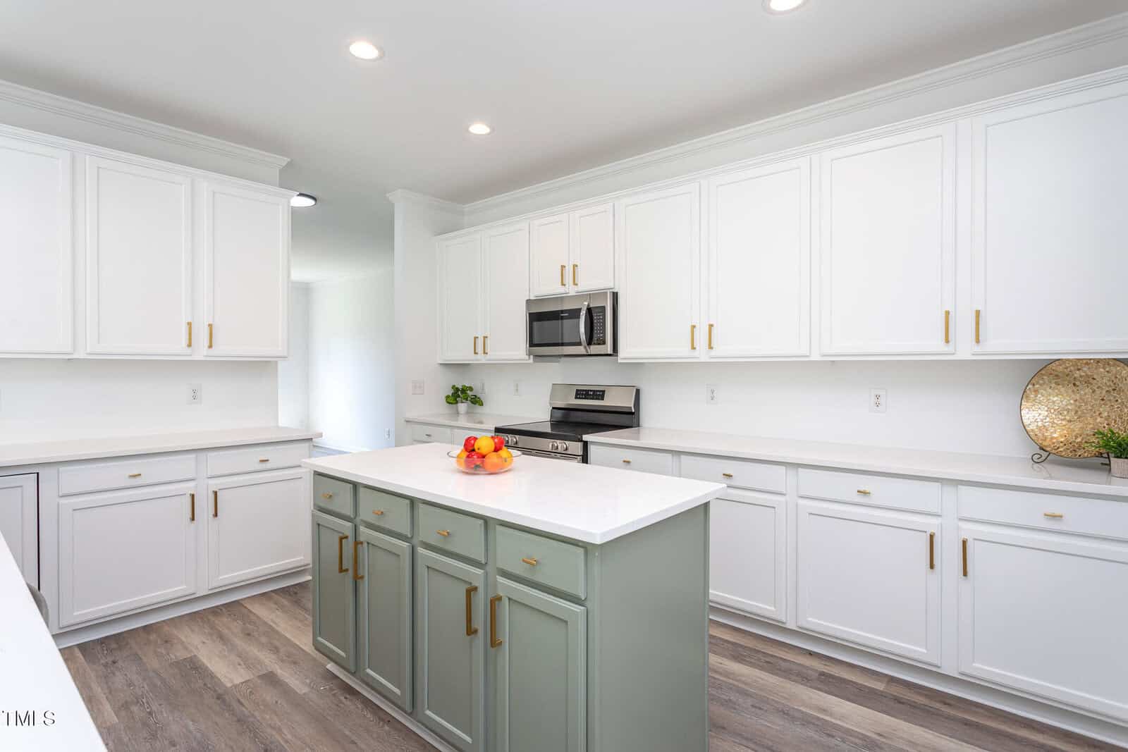 Modern kitchen with white cabinets, gold handles, a stainless steel stove and microwave, and a green island with a white countertop. A bowl of fruit and a decorative plate are on display. Wood-style flooring throughout.