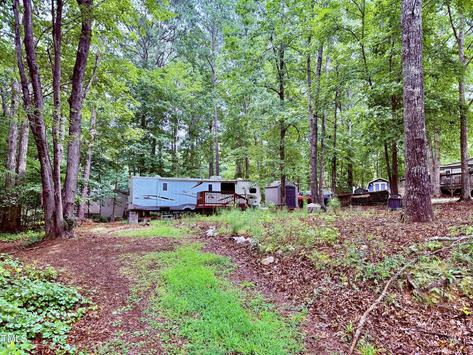 A large RV is parked in a wooded area with green grass and fallen leaves at 110 Arapaho Drive, Louisburg. There is a small deck attached to the RV and a shed nearby. Several trees surround the scene, with other trailers visible in the background.