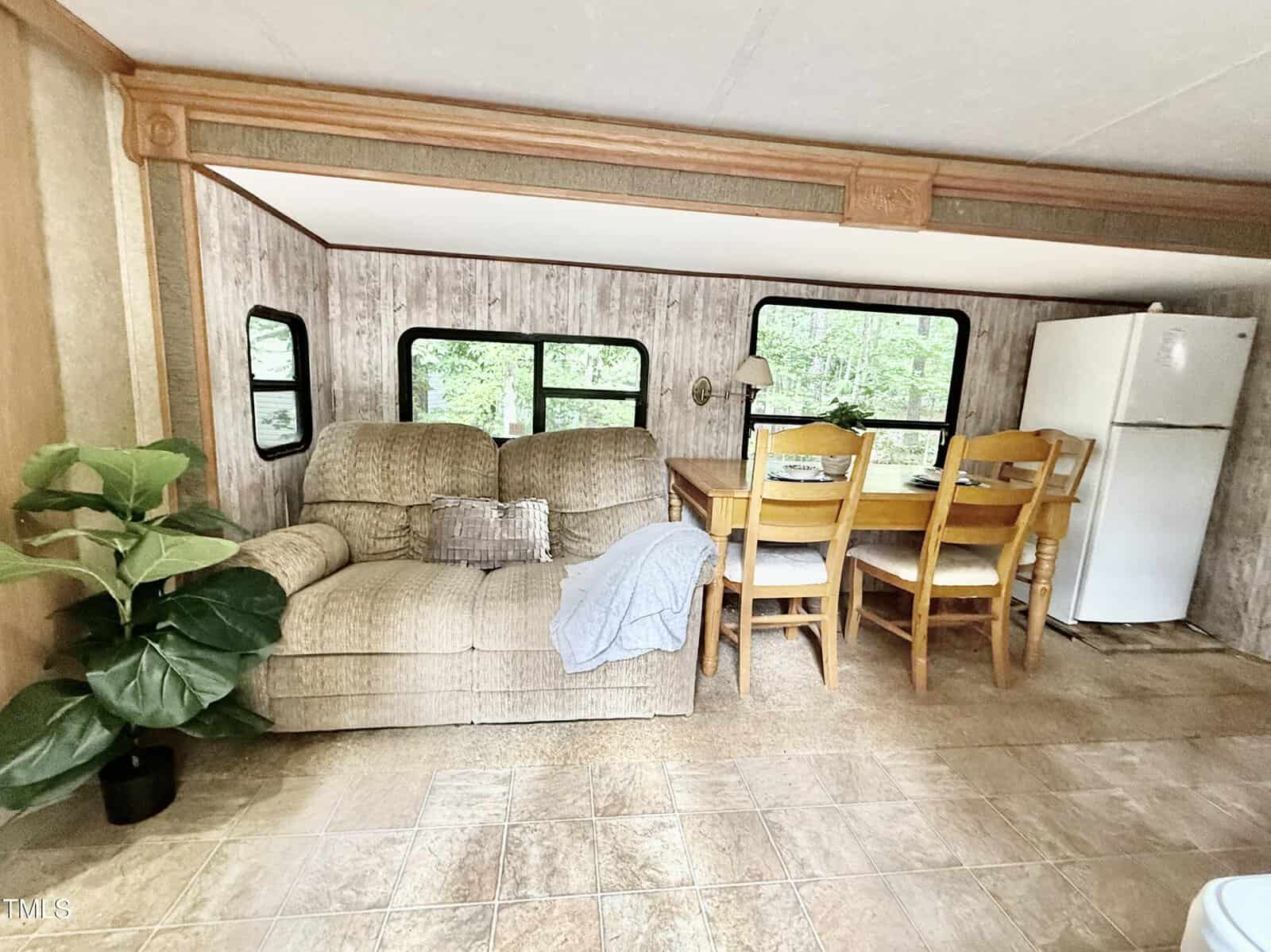 A beige loveseat with a pillow and blanket sits next to a potted plant. Nearby is a wooden dining table with four chairs, a lamp, and a white refrigerator. Large windows show trees outside. The floor is tiled.