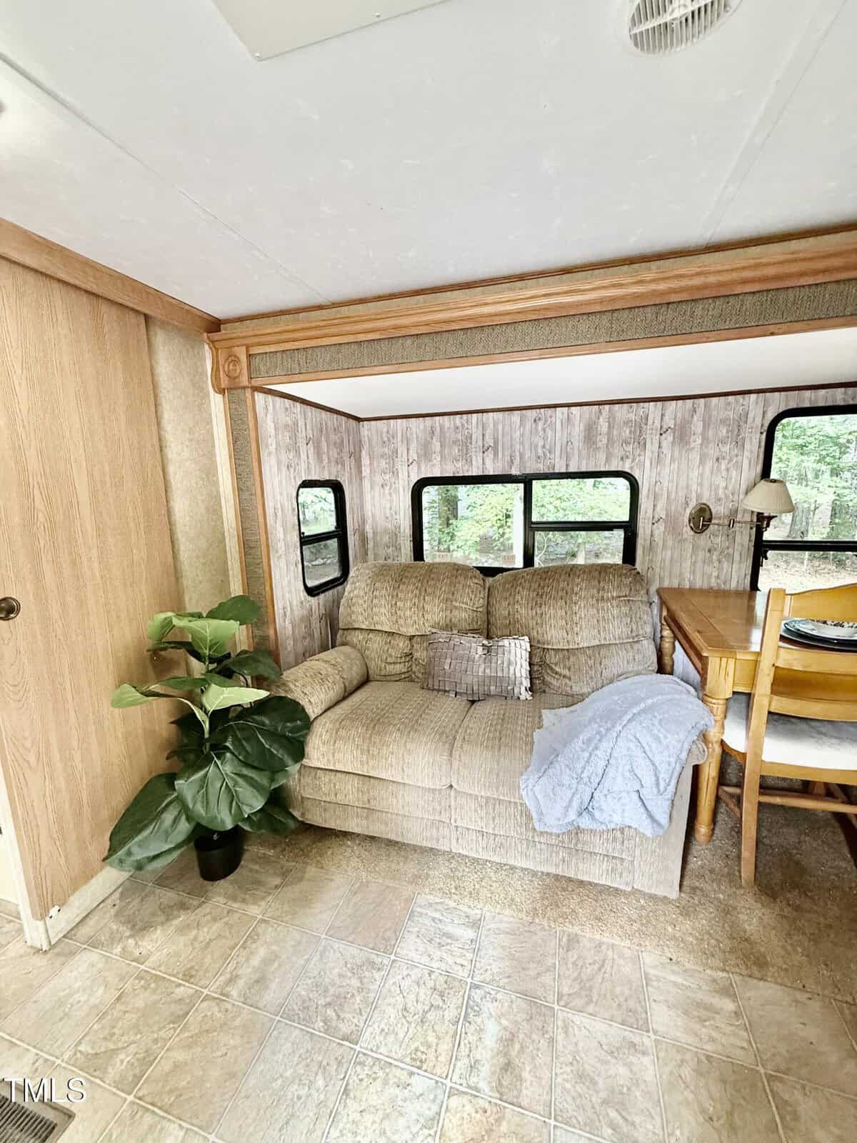 A small living area with a beige loveseat, silver pillow, blue blanket, wooden dining table with chairs, potted plant, tile floor, wood-paneled walls, and several windows showing trees outside.