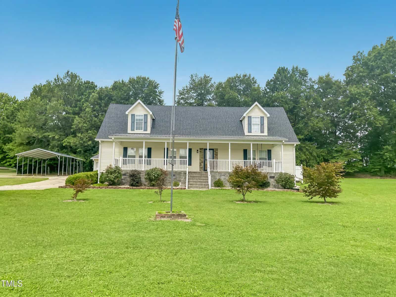 A two-story house at 920 Vanco Mill Road in Henderson features light-colored siding, a dark roof, and dormer windows. It sits on a large, well-maintained lawn with small shrubs, an American flag out front, and trees lining the background.
