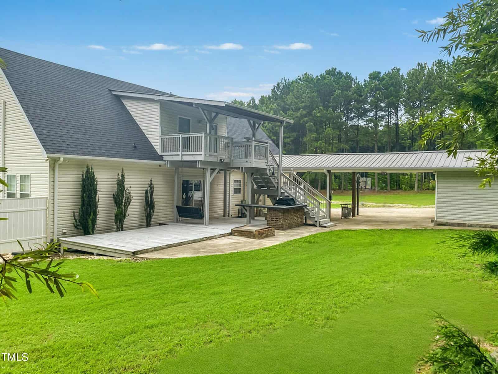 A two-story beige house with a covered second-floor balcony, large wooden deck, outdoor bar area, detached garage, and green lawn bordered by trees on a clear day.