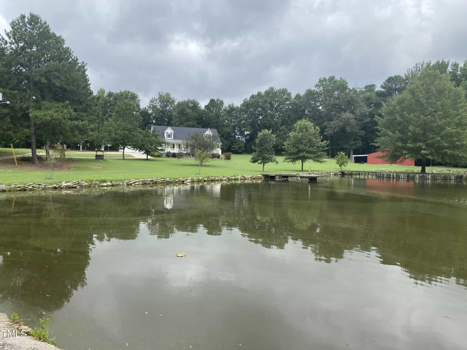 A pond with still water reflecting trees and a cloudy sky, with a grassy lawn, a white house, and a red barn-like building in the background. A small dock extends over the water near the trees.