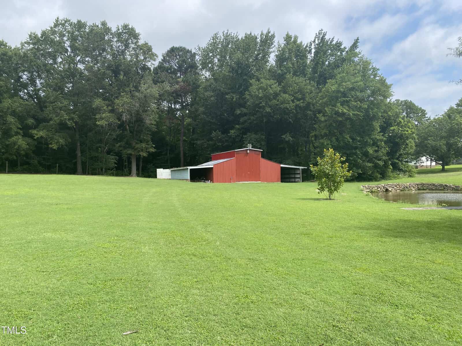 A red barn with a white roof sits on a large, grassy lawn. Trees line the background, and there is a small pond to the right of the barn. The sky is partly cloudy.
