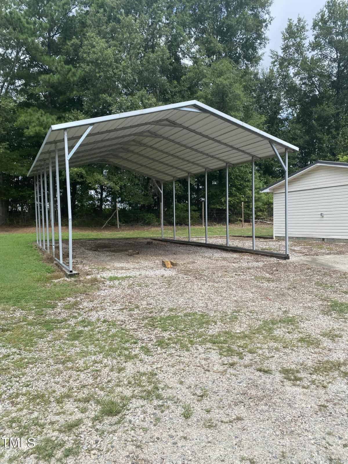 A metal carport with a peaked roof and open sides stands on a gravel area next to a light-colored building, surrounded by grass and trees.