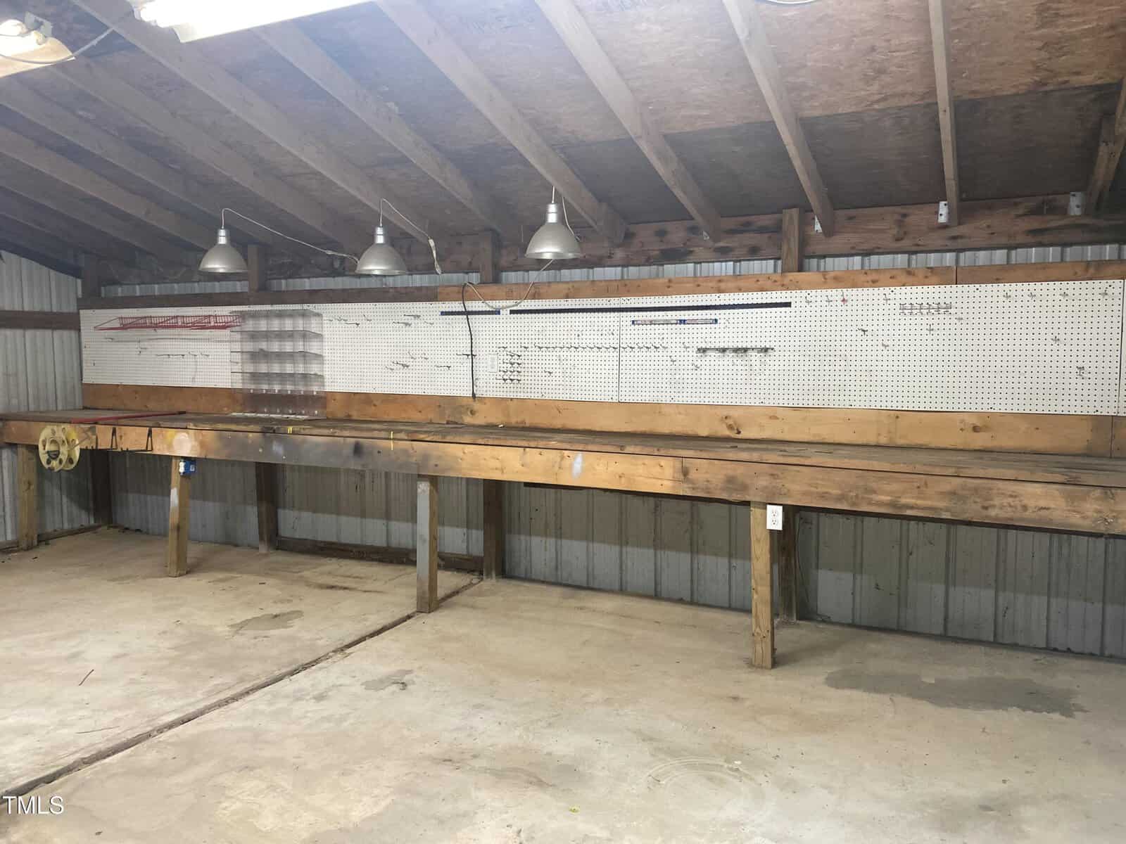 Wooden workbench under a sloped roof with hanging lights above. Pegboard with various hooks, rods, and small storage bins is mounted on the wall behind the bench. Concrete floor and exposed beams visible.