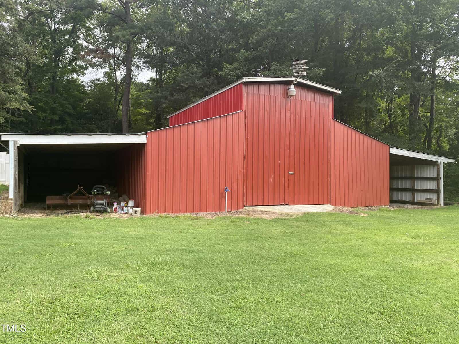 A red metal barn with two open, covered side areas stands on green grass, with trees in the background. Various equipment and cans are stored under the left side area.